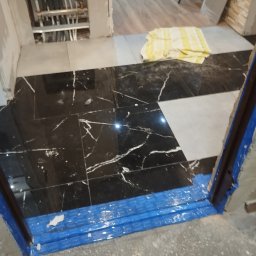 REMO DESIGN - Ułożona podłoga z czarno-białych płytek w trakcie prac wykończeniowych. Widoczne rury ogrzewania podłogowego i zabezpieczone folią drzwi. W tle odkurzacz.