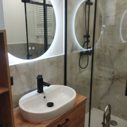 REMO DESIGN - Nowoczesna łazienka z owalną umywalką na drewnianej szafce, czarną armaturą i podświetlanym lustrem. Kabina prysznicowa z czarnymi ramami i marmurowe płytki.