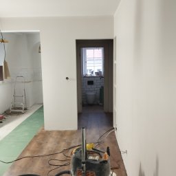 REMO DESIGN - Wnętrze po malowaniu: białe ściany, podłoga z paneli, widok na wyremontowaną łazienkę z białymi płytkami. Na pierwszym planie odkurzacz przemysłowy.