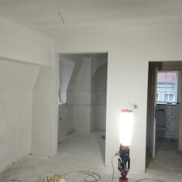 REMO DESIGN - Wnętrze po szpachlowaniu i malowaniu na biało, widoczny agregat malarski i lampa budowlana. W tle wykończona łazienka z białymi płytkami i toaletą.