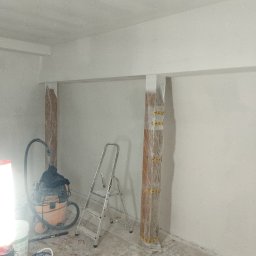 REMO DESIGN - Przygotowanie do malowania: zabezpieczone folią kolumny, drabina, odkurzacz i narzędzia malarskie w pomieszczeniu z świeżo pomalowanymi ścianami na biało.