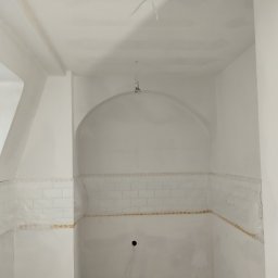 REMO DESIGN - Biała łazienka w trakcie remontu: częściowo ułożone białe płytki, ściany przygotowane do malowania, widoczne kable oświetleniowe. Łuk nad fragmentem ściany z płytkami.