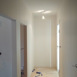 REMO DESIGN - Jasny korytarz po malowaniu ścian na biało. Na podłodze płyty OSB i resztki taśmy malarskiej. Widoczne drzwi i fragmenty innych pomieszczeń. Oświetlenie sufitowe.