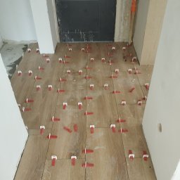 REMO DESIGN - Układanie drewnopodobnych płytek podłogowych z użyciem czerwonych klipsów poziomujących, widok z góry na świeżo ułożoną podłogę w korytarzu, ciemne drzwi w tle.