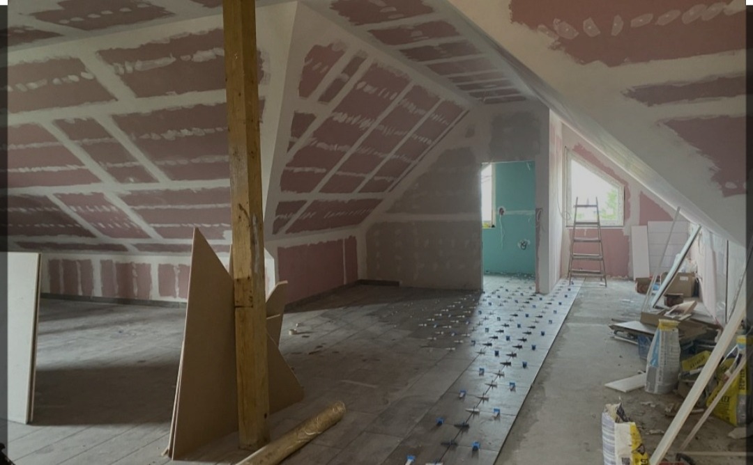 Poddasze w trakcie prac wykończeniowych: ułożona częściowo podłoga z płytek z krzyżykami dystansowymi, ściany i skośny sufit przygotowane pod gładzie, widoczna drabina i materiały budowlane.