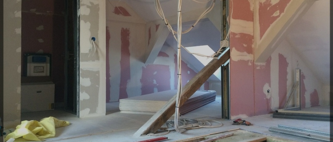 Poddasze w trakcie adaptacji: widoczne płyty karton-gips, drewniana belka wzmacniająca, przewody elektryczne i narzędzia na podłodze. Ściany przygotowane do dalszych prac wykończeniowych.