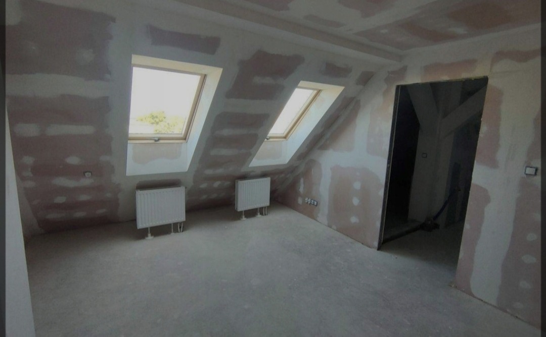 Poddasze w trakcie prac wykończeniowych: ściany pokryte warstwą gładzi szpachlowej, dwa okna dachowe, grzejniki zamontowane, widoczne ślady po instalacjach elektrycznych.