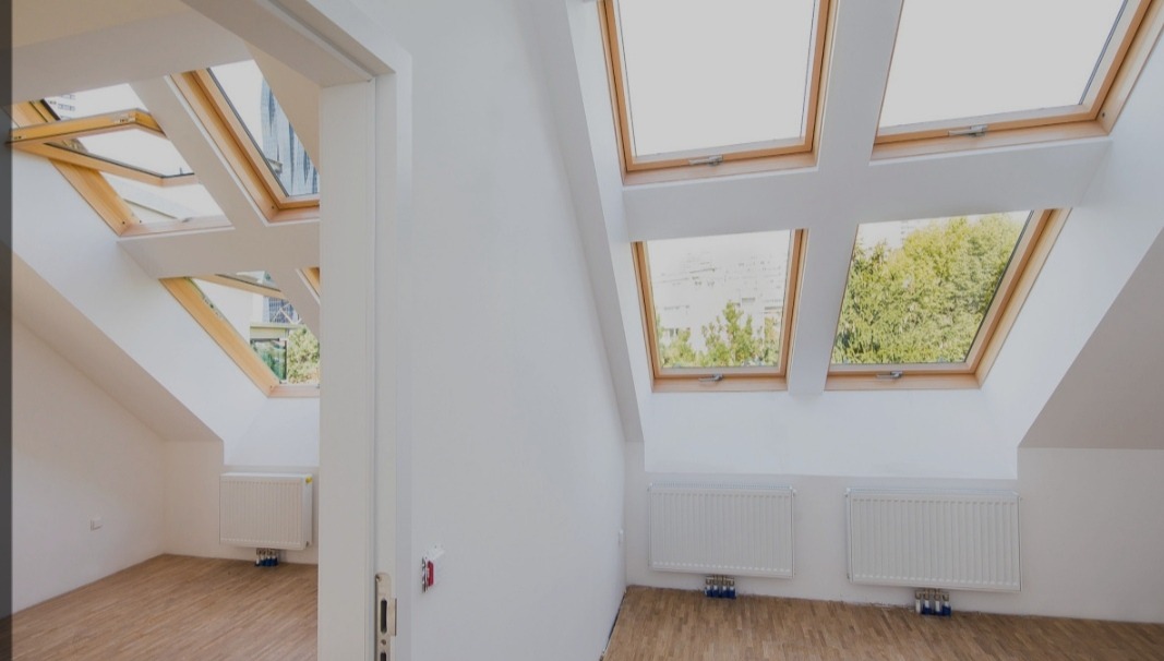 Jasne poddasze z drewnianymi oknami dachowymi, białymi ścianami, grzejnikami i parkietem, widok z otwartych drzwi do drugiego pokoju.