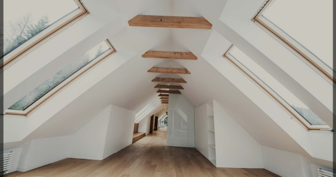 Jasne poddasze z drewnianą podłogą, skośnymi ścianami, oknami dachowymi z widokiem na drzewa oraz dekoracyjnymi belkami stropowymi