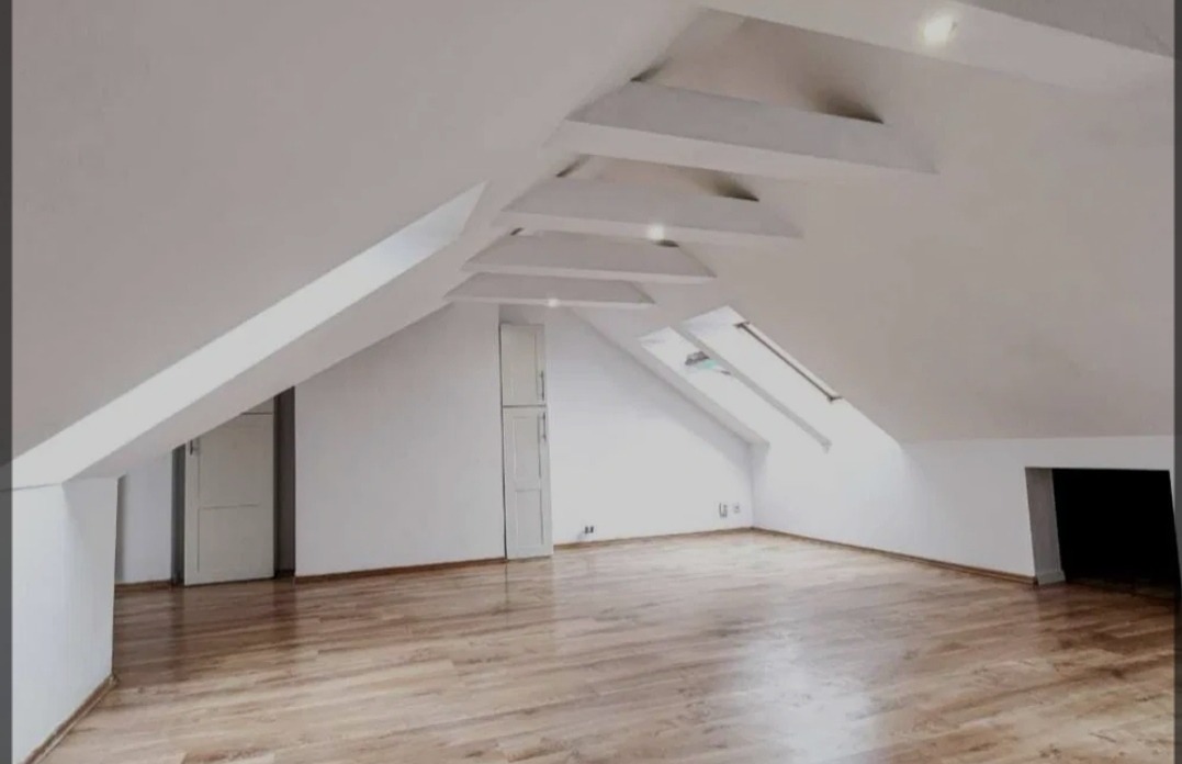 Poddasze z białymi ścianami, drewnianą podłogą, oknami dachowymi i dekoracyjnymi elementami sufitowymi.