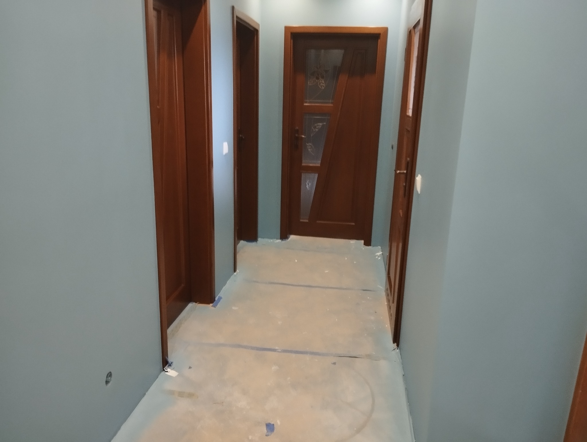 Korytarz w trakcie malowania, z trzema drewnianymi drzwiami, podłoga zabezpieczona folią malarską, ściany pomalowane na szaro-niebieski kolor.
