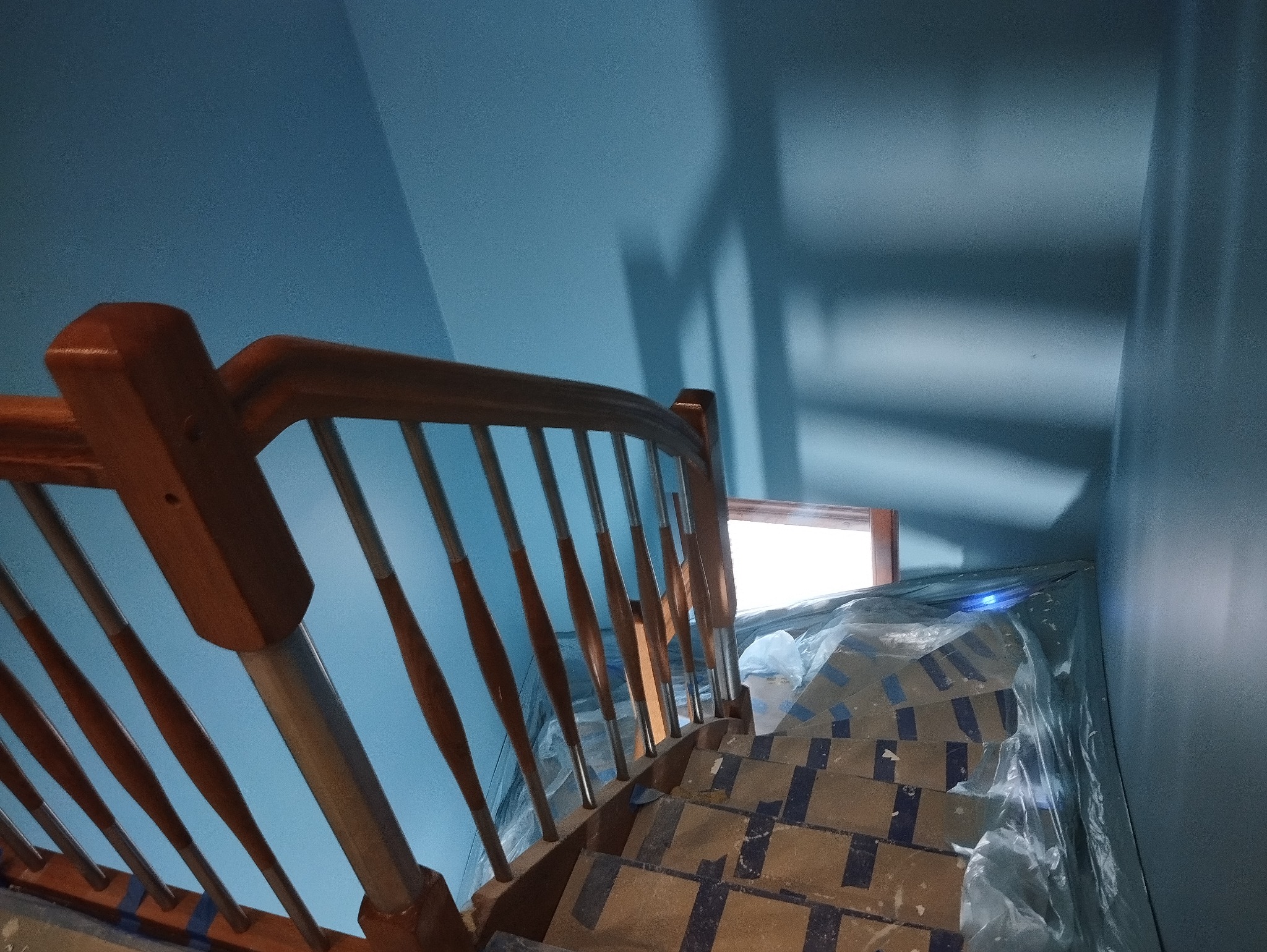 Schody zabezpieczone kartonem i folią malarską podczas prac wykończeniowych, drewniana balustrada z metalowymi tralkami, ściany pomalowane na niebiesko.