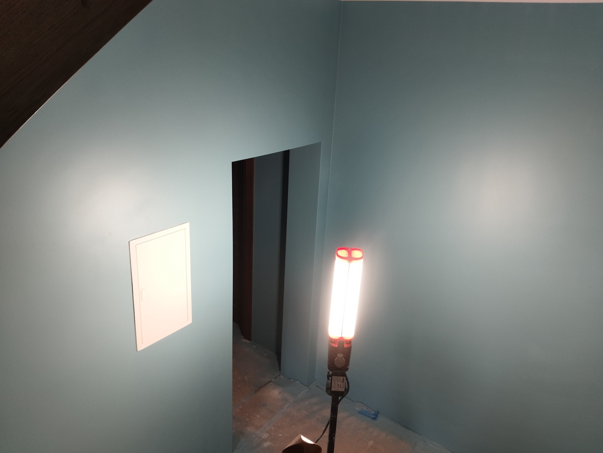 Świeżo pomalowane na niebiesko ściany w pomieszczeniu z widocznym wejściem do innego pokoju, oświetlone lampą roboczą, z klapką rewizyjną na ścianie.