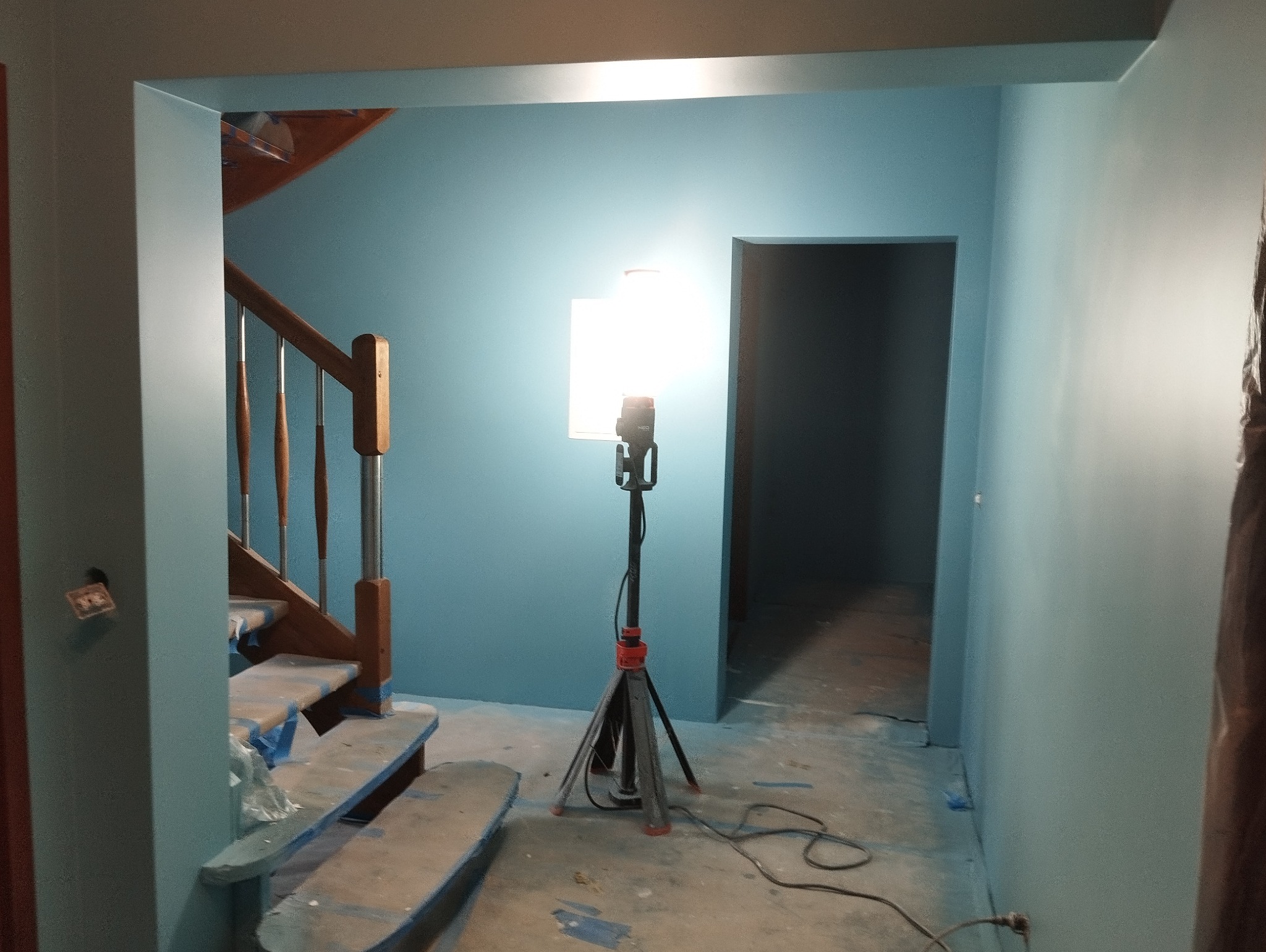 Świeżo pomalowane na niebiesko ściany w Krosno Odrzańskie, widoczne schody zabezpieczone folią malarską oraz lampa budowlana na trójnogu oświetlająca wnętrze.