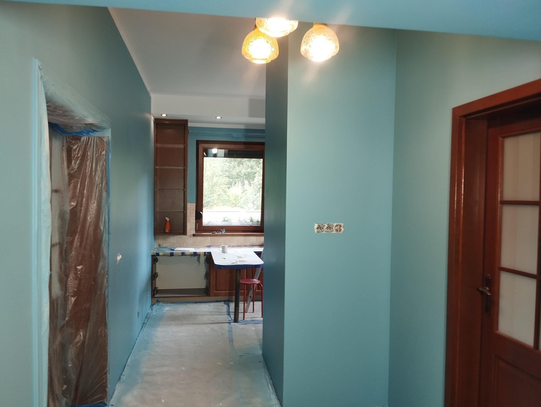 Turkusowe ściany po malowaniu w trakcie remontu, zabezpieczone folią drzwi, widoczne gniazdka elektryczne i fragment kuchni w tle.