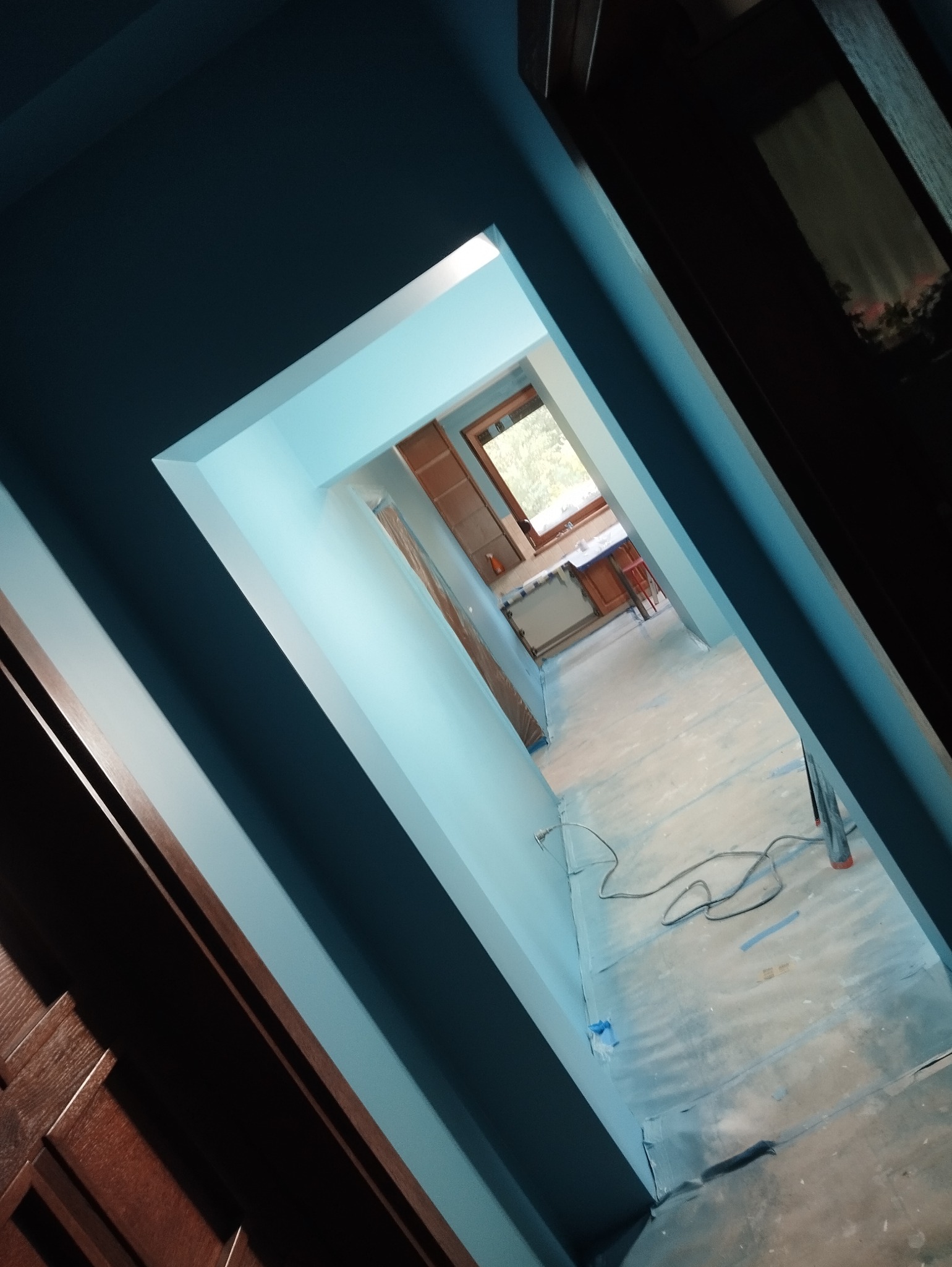 Wnętrze mieszkania w trakcie malowania, widok z korytarza na kuchnię, ściany pomalowane na jasny odcień błękitu, podłoga zabezpieczona folią malarską, widoczne kable i drabina.