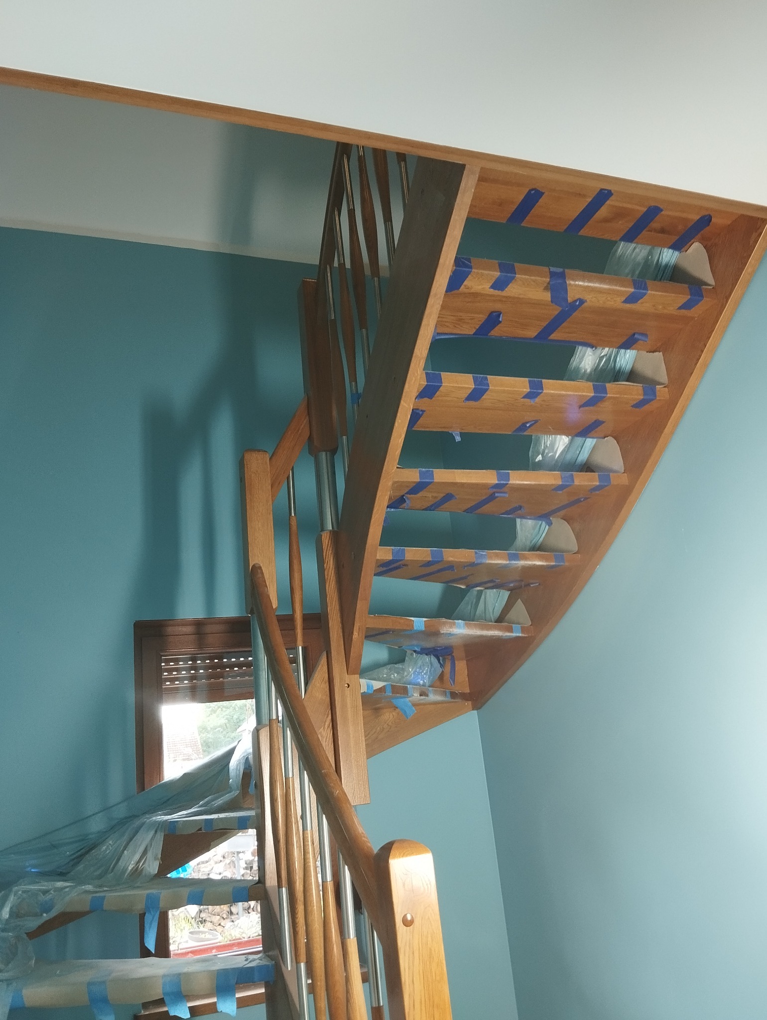 Drewniane schody z metalowymi balustradami, zabezpieczone niebieską taśmą malarską i folią ochronną, podczas prac wykończeniowych, na tle turkusowej ściany.