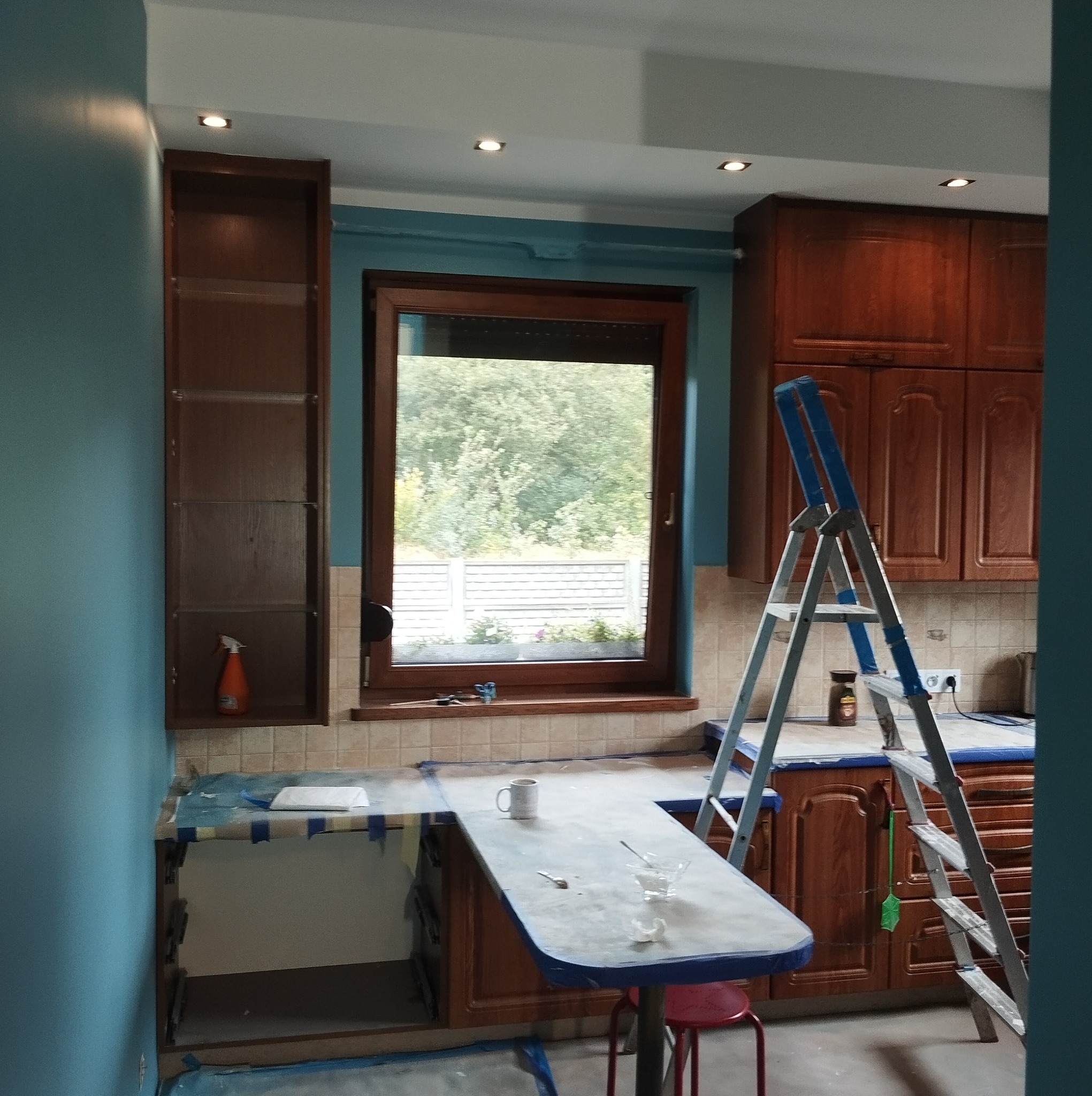 Kuchnia w trakcie malowania ścian na kolor turkusowy, zabezpieczone blaty taśmą malarską, drabina aluminiowa oparta o szafki, widoczne narzędzia i akcesoria malarskie.