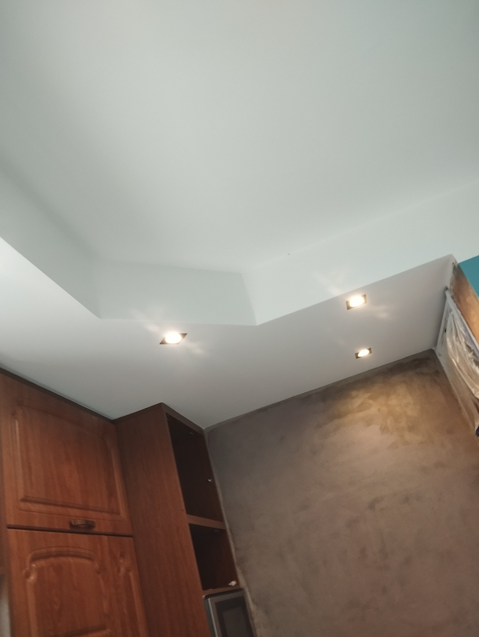 Sufit podwieszany z oświetleniem punktowym w trakcie prac wykończeniowych, widoczne drewniane szafki kuchenne i fragment ściany z nałożoną strukturą.