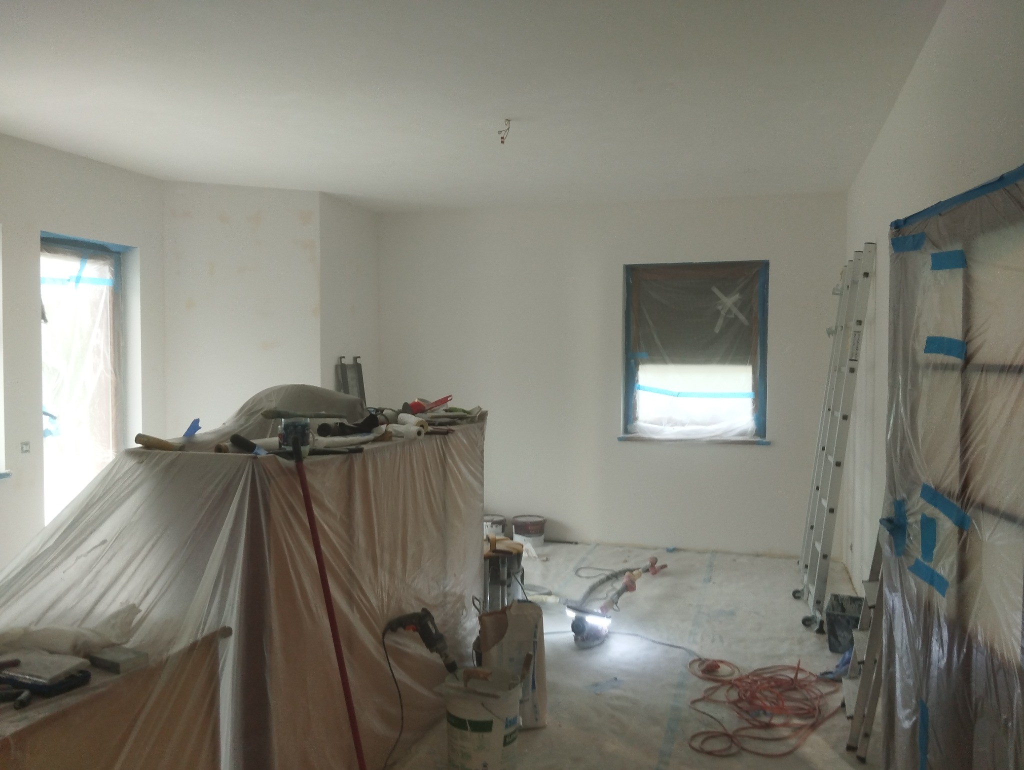 Pokój w trakcie malowania: zabezpieczone okna i drzwi folią, rozłożona folia ochronna na podłodze, drabina oparta o ścianę, narzędzia malarskie przykryte folią, widoczne ślady po szpachlowaniu...