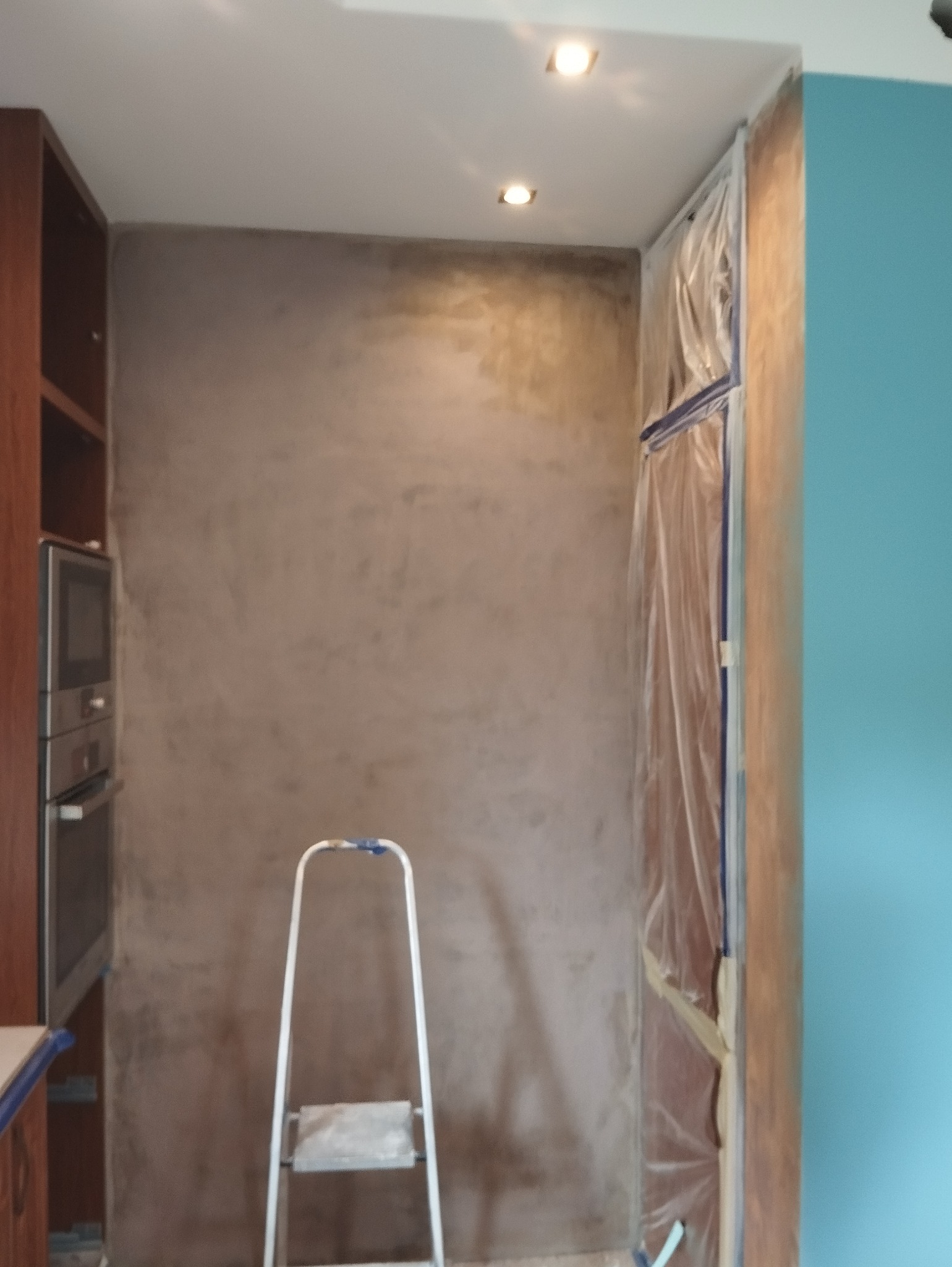 Ściana w trakcie wykańczania z nałożoną gładzią w odcieniach beżu, zabezpieczone folią meble i drabina aluminiowa w pomieszczeniu z oświetleniem punktowym.