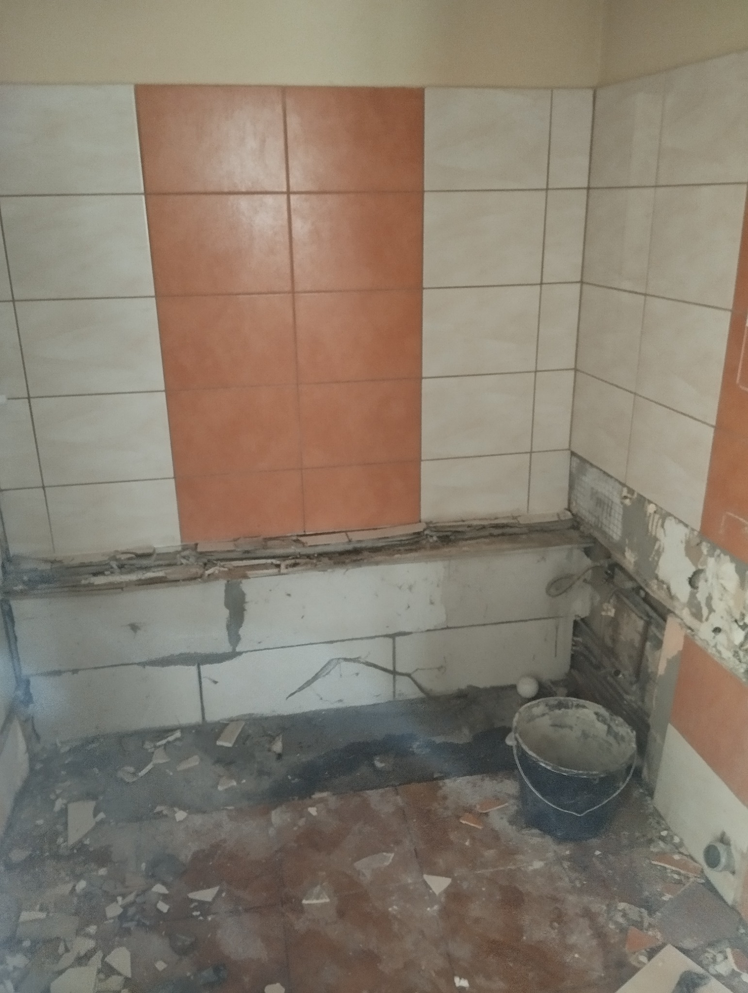 Demontaż płytek w łazience, widoczne uszkodzone ściany, gruz na podłodze i wiadro zaprawy, fragment ściany z białymi i pomarańczowymi płytkami
