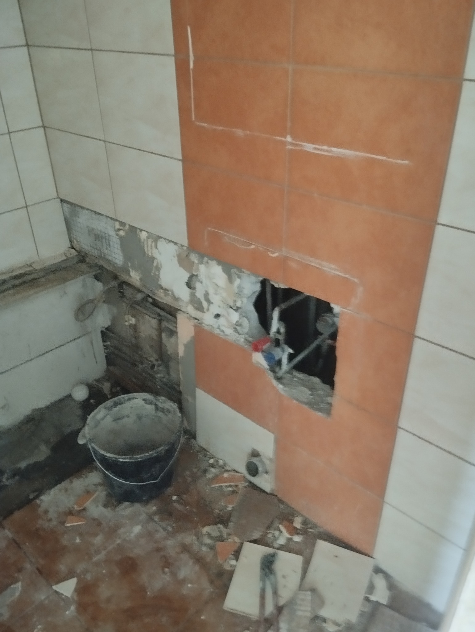 Demontaż płytek w łazience, odsłonięte rury wodne, widoczne ślady po kleju, fragmenty płytek na podłodze, czarne wiadro z resztkami zaprawy.