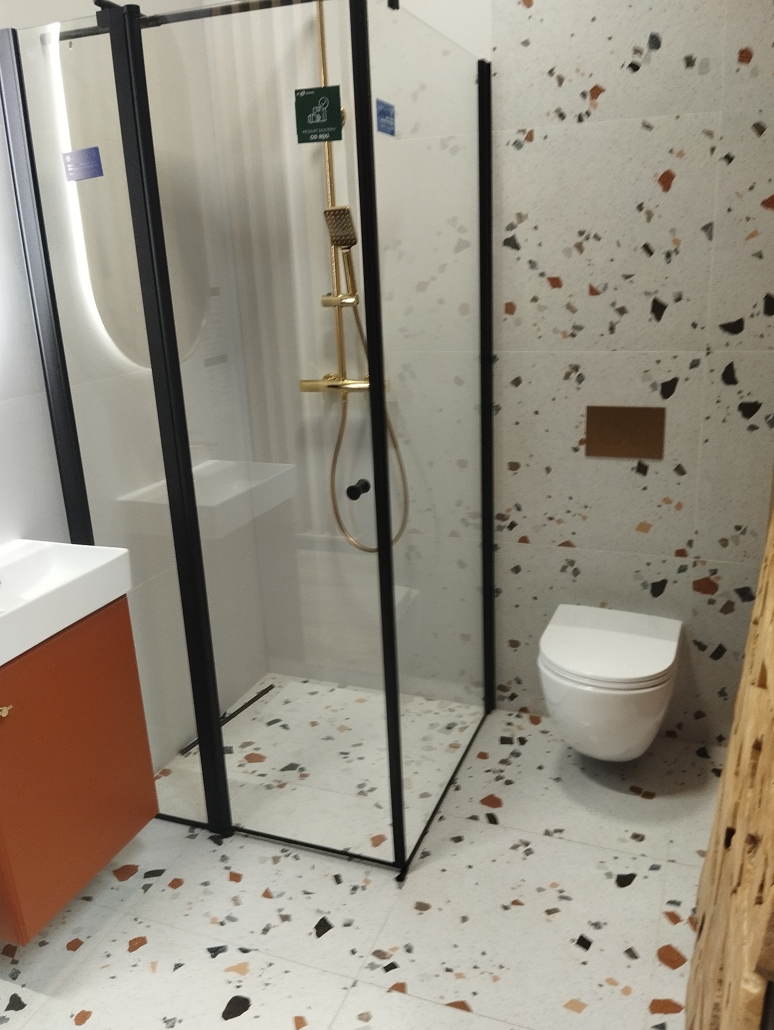 Nowoczesna łazienka z kabiną prysznicową z czarną ramą, umywalką na pomarańczowej szafce i toaletą, wykończona płytkami imitującymi lastryko.