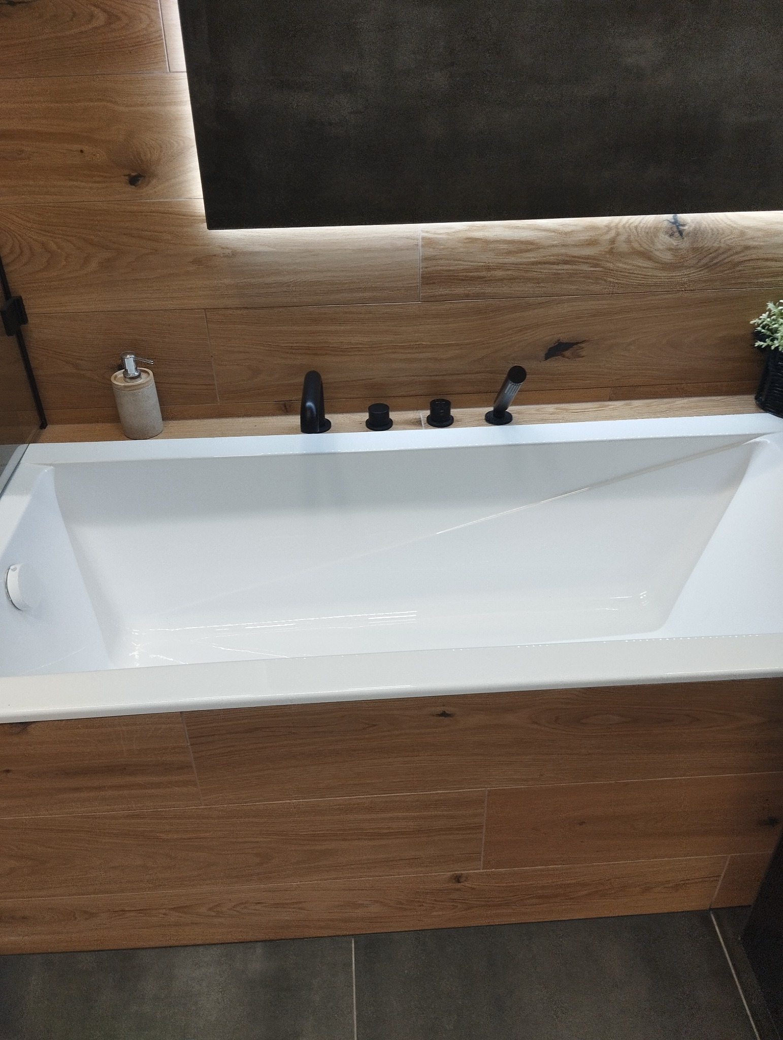 Nowoczesna biała wanna obudowana płytkami imitującymi drewno, czarna armatura, podświetlane lustro i szara podłoga.