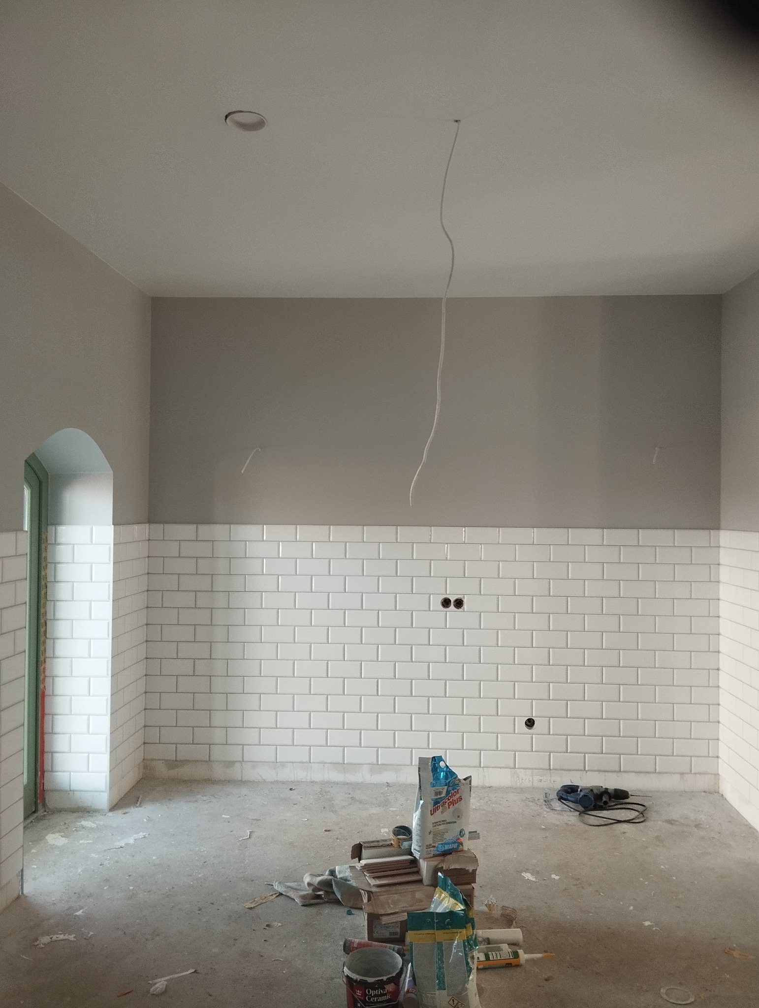 Łazienka w trakcie remontu: białe, prostokątne płytki ułożone do połowy ściany, szare ściany powyżej, widoczne narzędzia i materiały budowlane na podłodze. W tle łukowe wejście.