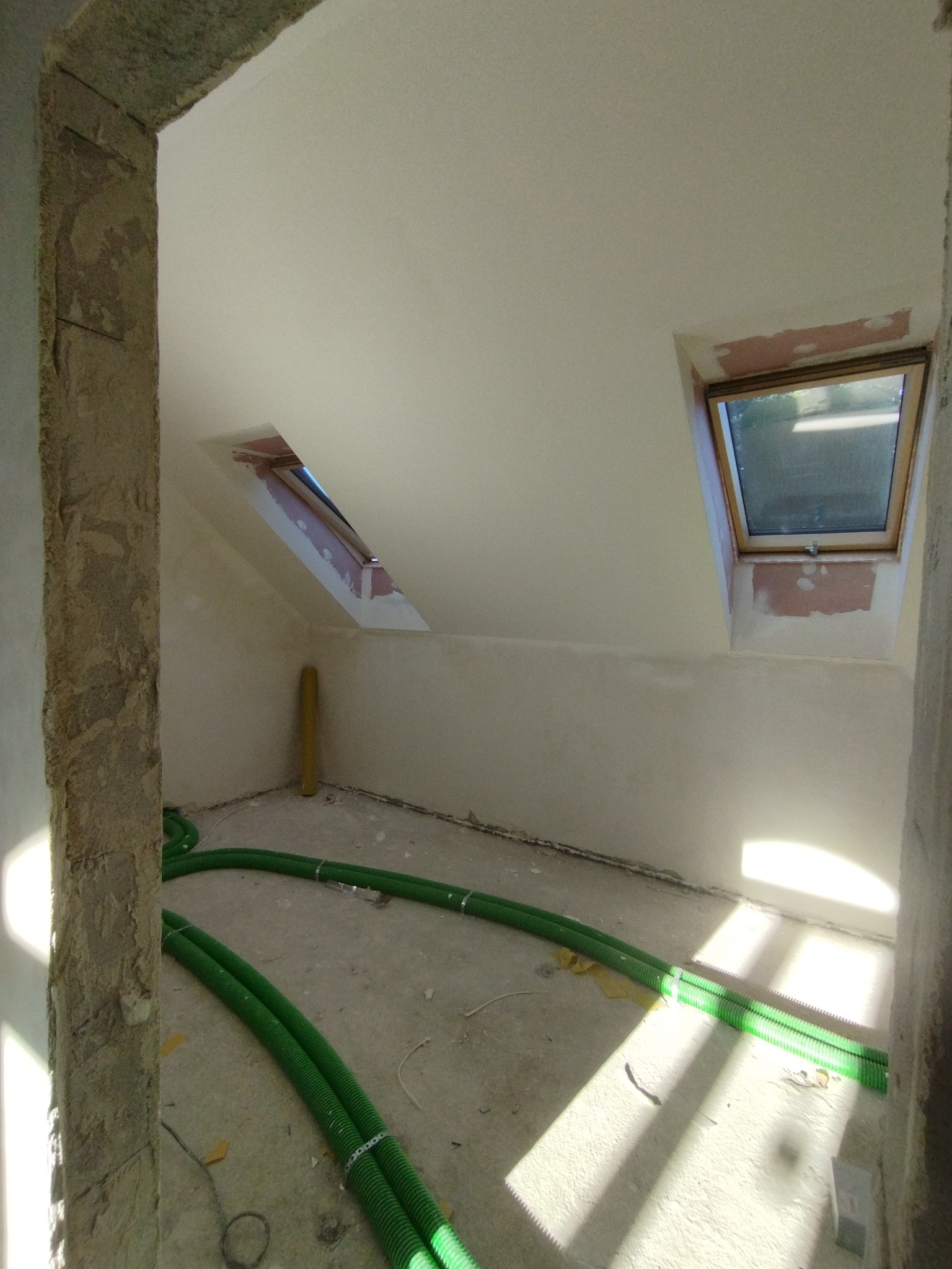 Poddasze po gładziowaniu, widoczne okna dachowe i zielone rury wentylacyjne na podłodze. Ściany i sufit w jasnym odcieniu bieli, surowy stan wykończenia.