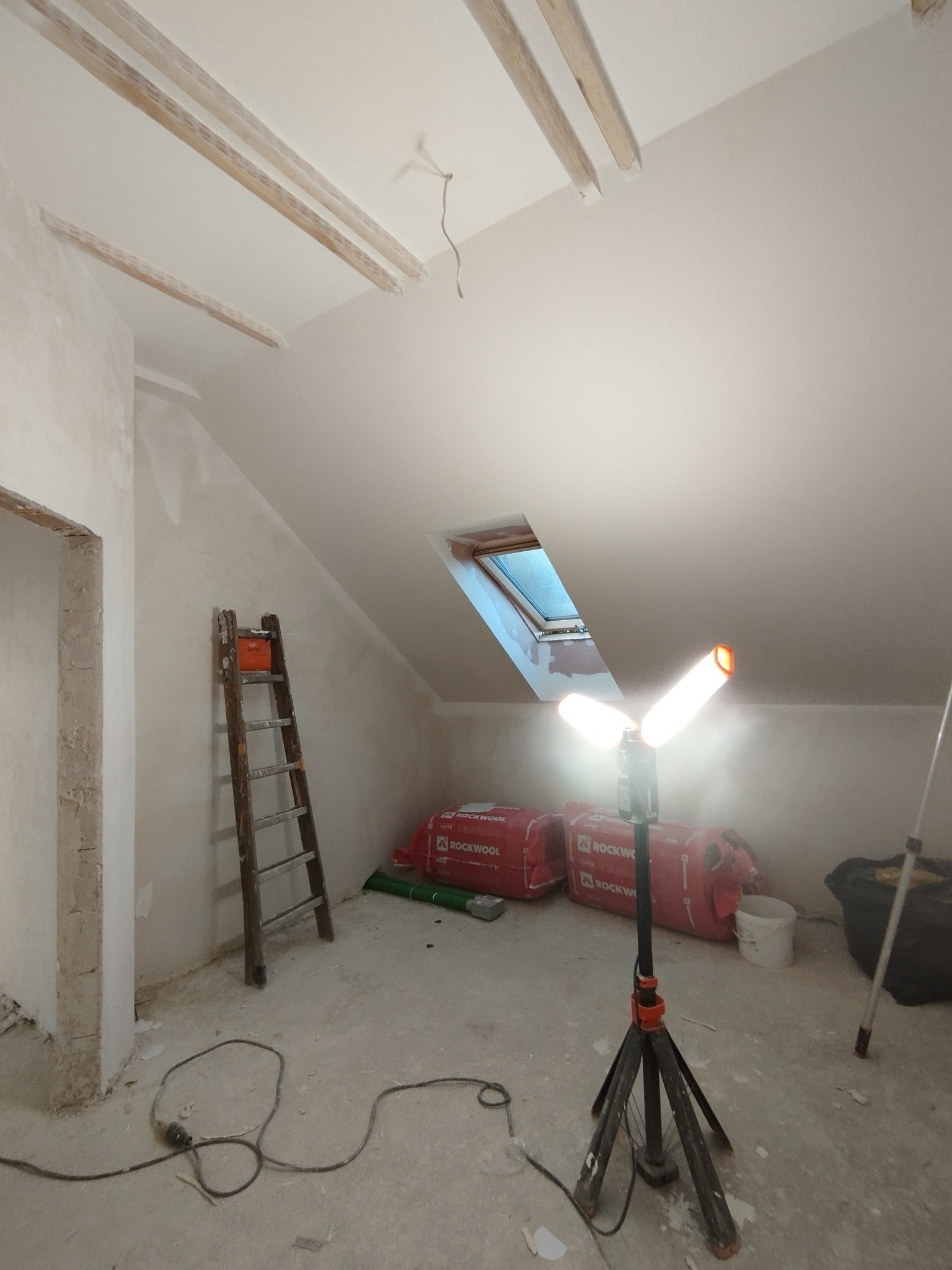 Poddasze w trakcie wykańczania: gładzie na ścianach, drewniane belki na suficie, okno dachowe, oświetlenie budowlane i materiały izolacyjne Rockwool.