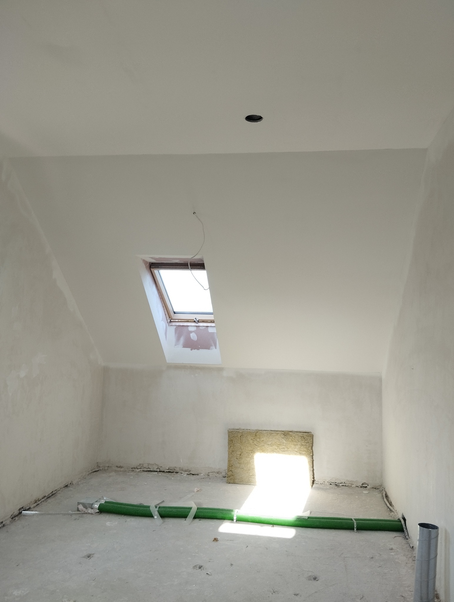 Poddasze w trakcie prac wykończeniowych: gładzie na ścianach, okno dachowe, widoczne przewody wentylacyjne na podłodze, przygotowane miejsce na kominek.