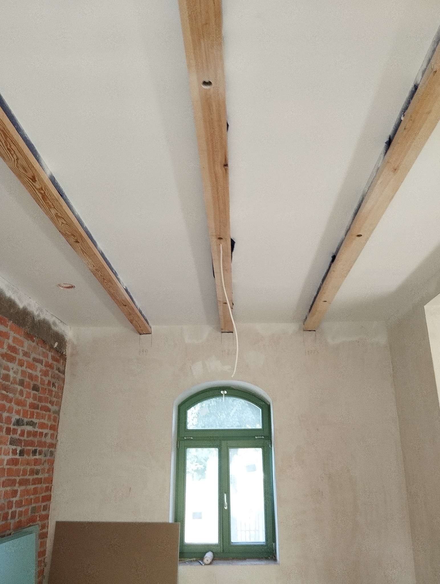 Wnętrze w trakcie remontu: surowa cegła, okno w zielonej ramie, drewniane belki stropowe z instalacją elektryczną, ściany przygotowane pod gładzie.