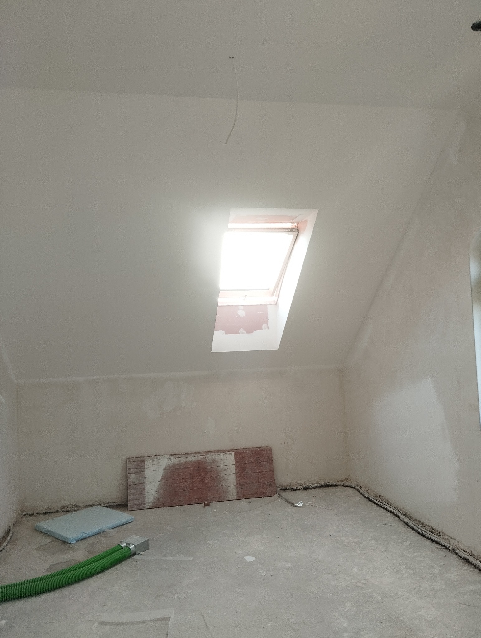 Pokój na poddaszu w trakcie prac wykończeniowych. Widoczne okno dachowe, ściany przygotowane do malowania, podłoga z elementami instalacji i zabezpieczeń.