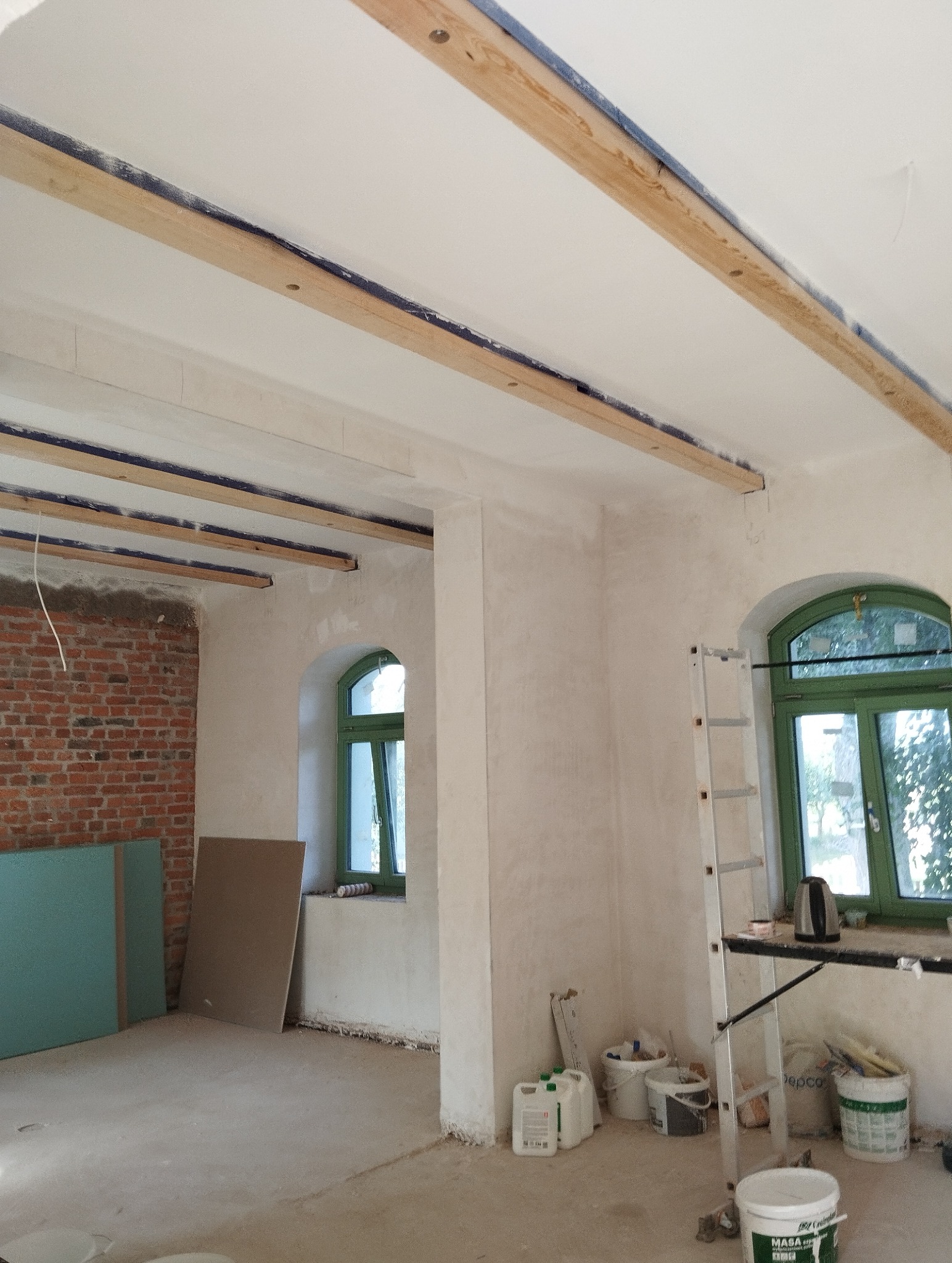 Wnętrze w trakcie remontu: surowe ściany po gładzi, drewniane belki pod sufitem z niebieską taśmą, okna z zielonymi ramami, drabina i materiały budowlane.