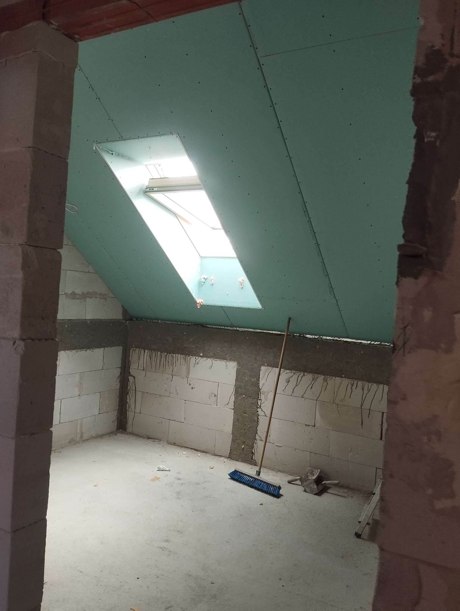 Poddasze w trakcie adaptacji: ściany z bloczków, sufit z płyt karton-gips z oknem dachowym. Widoczne narzędzia i szczotka. Surowy stan wykończenia.