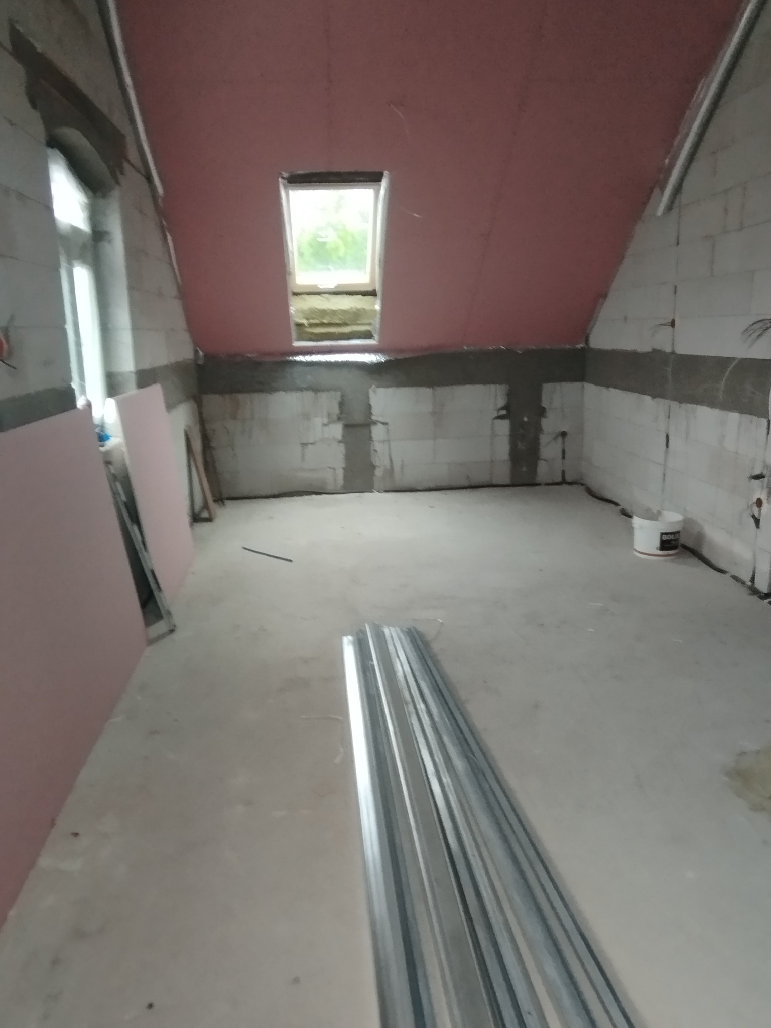 Poddasze w trakcie adaptacji: ściany z bloków, okno dachowe, płyty karton-gips i profile metalowe przygotowane do montażu. Widoczny etap prac wykończeniowych.