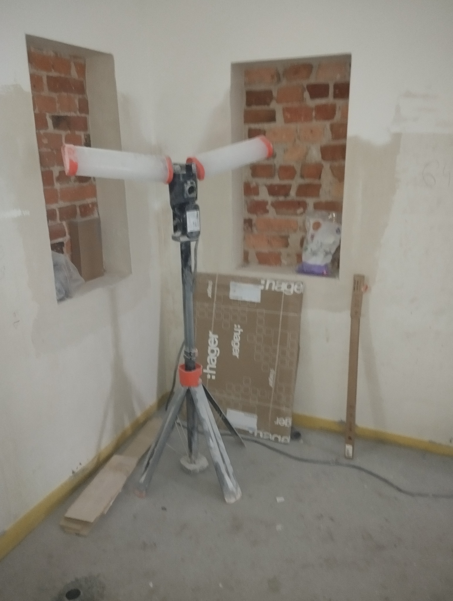 Wnętrze w trakcie prac wykończeniowych: ściany przygotowane do malowania, widoczne ceglane wnęki, lampa budowlana na statywie i kartonowe opakowania.