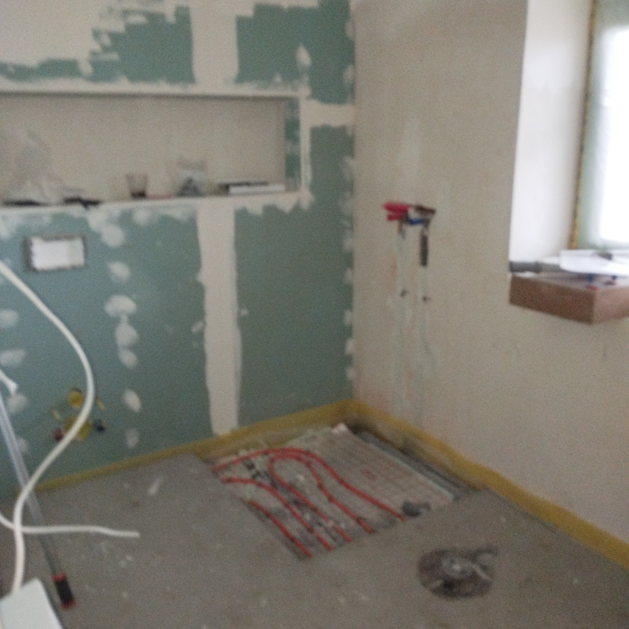 Łazienka w trakcie remontu: ściany z płyt gipsowo-kartonowych, instalacja ogrzewania podłogowego, widoczne rury i okno. Etap prac wykończeniowych.