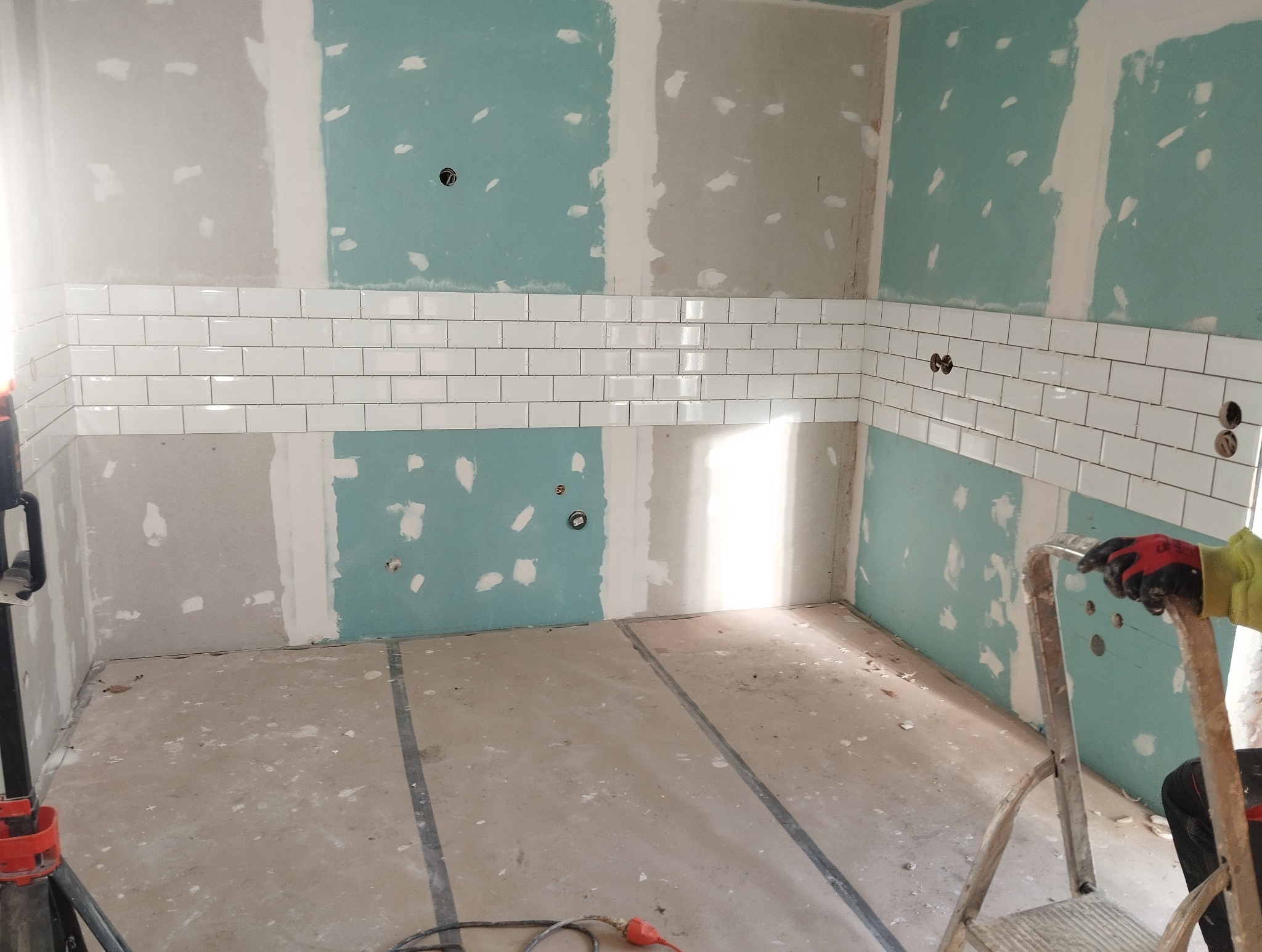 Wnętrze w trakcie prac wykończeniowych: białe, prostokątne płytki ułożone na ścianie, fragment drabiny i narzędzia na podłodze. Ściany częściowo pokryte płytą gipsową.