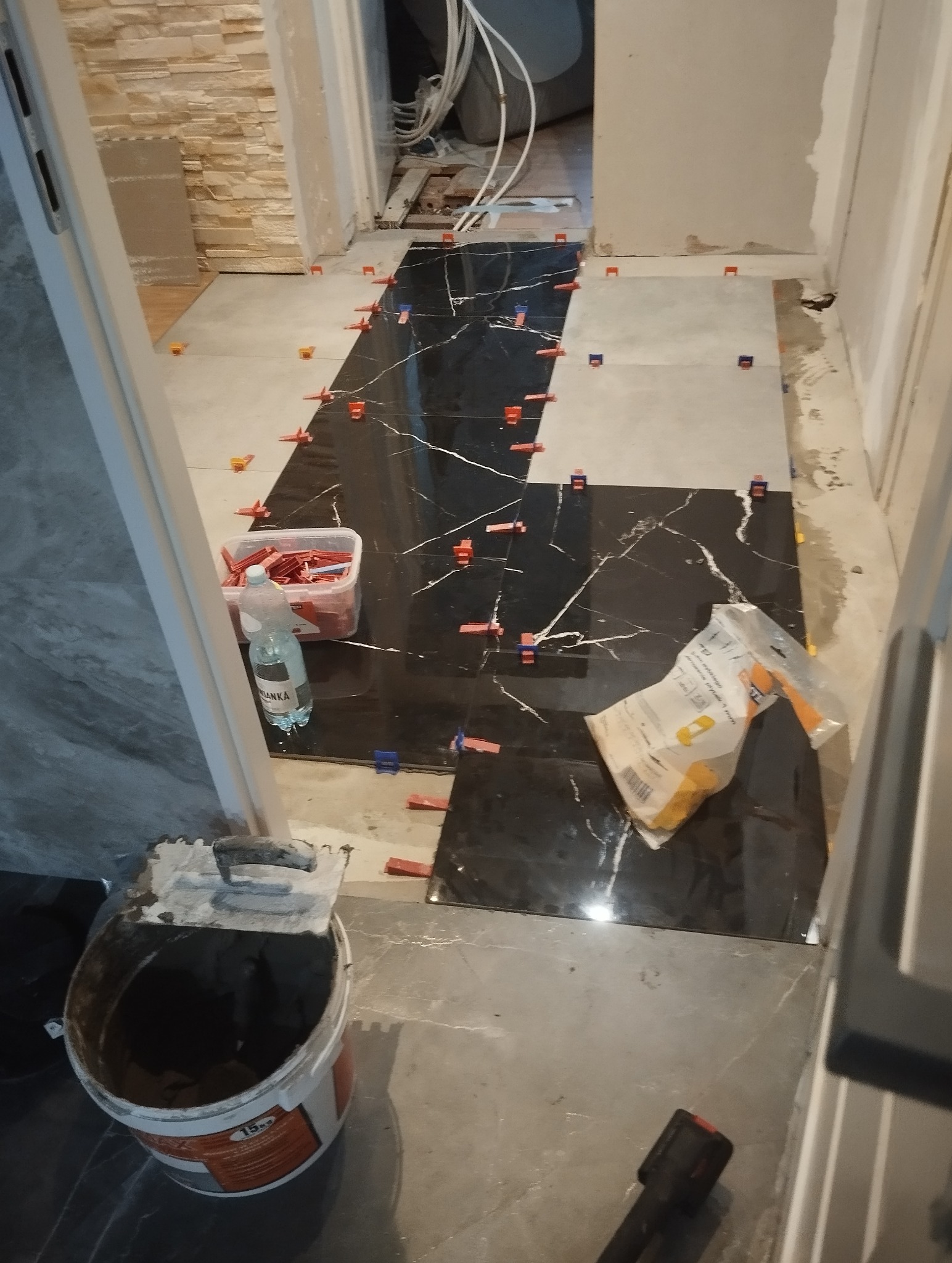 Układanie czarno-białych płytek podłogowych z marmurowym wzorem, z użyciem klipsów poziomujących. Widoczny kubeł z zaprawą i narzędzia glazurnicze.