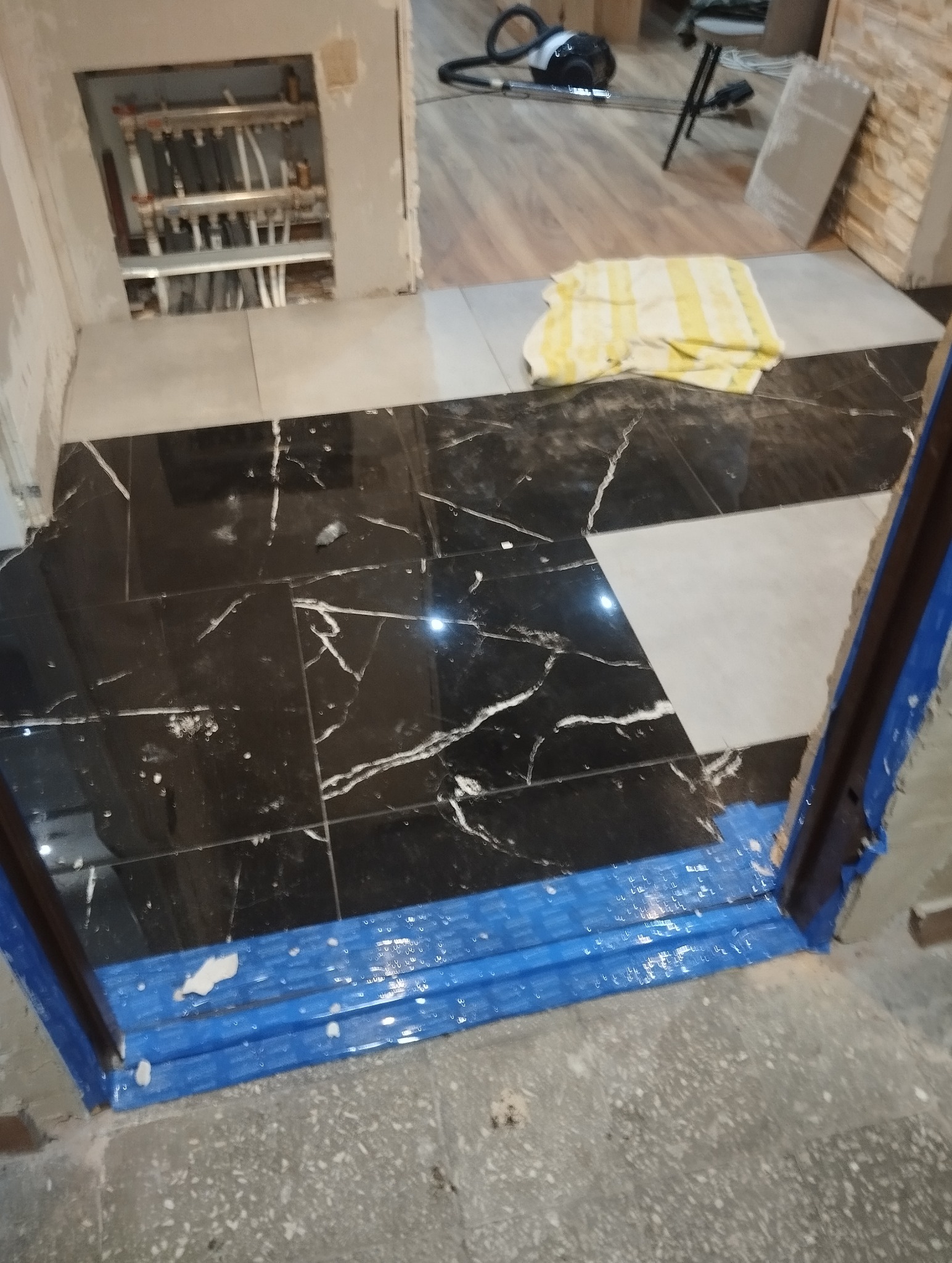 Ułożona podłoga z czarno-białych płytek w trakcie prac wykończeniowych. Widoczne rury ogrzewania podłogowego i zabezpieczone folią drzwi. W tle odkurzacz.