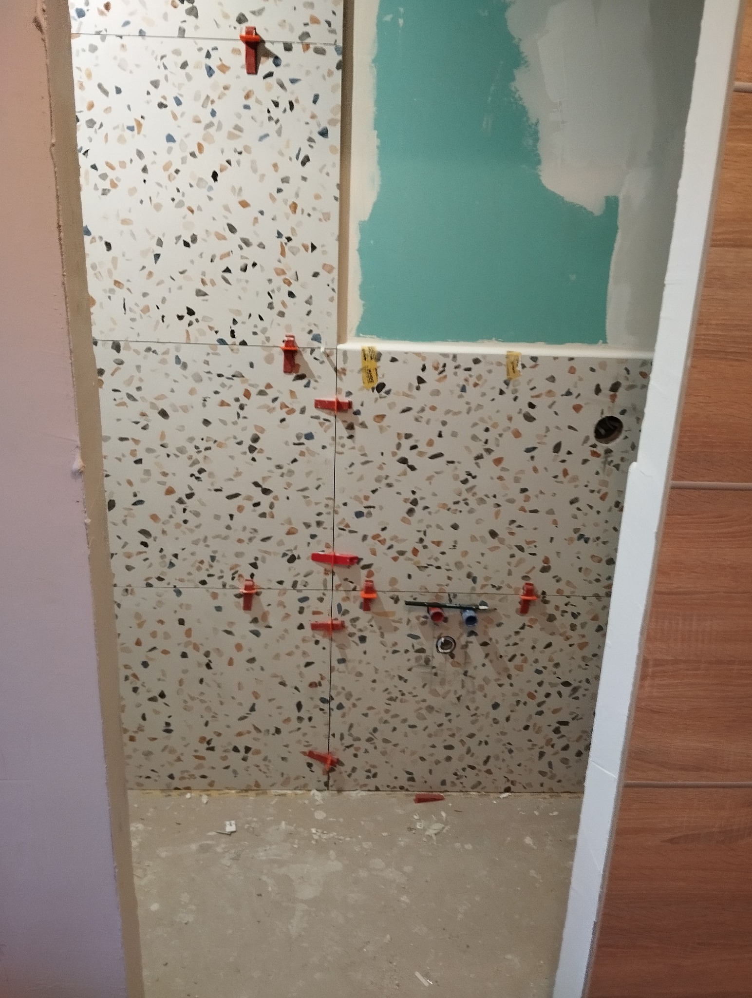 Układanie płytek lastryko w łazience z użyciem klipsów poziomujących. Widoczne fragmenty instalacji wodnej i pomalowana ściana. Prace glazurnicze w toku.