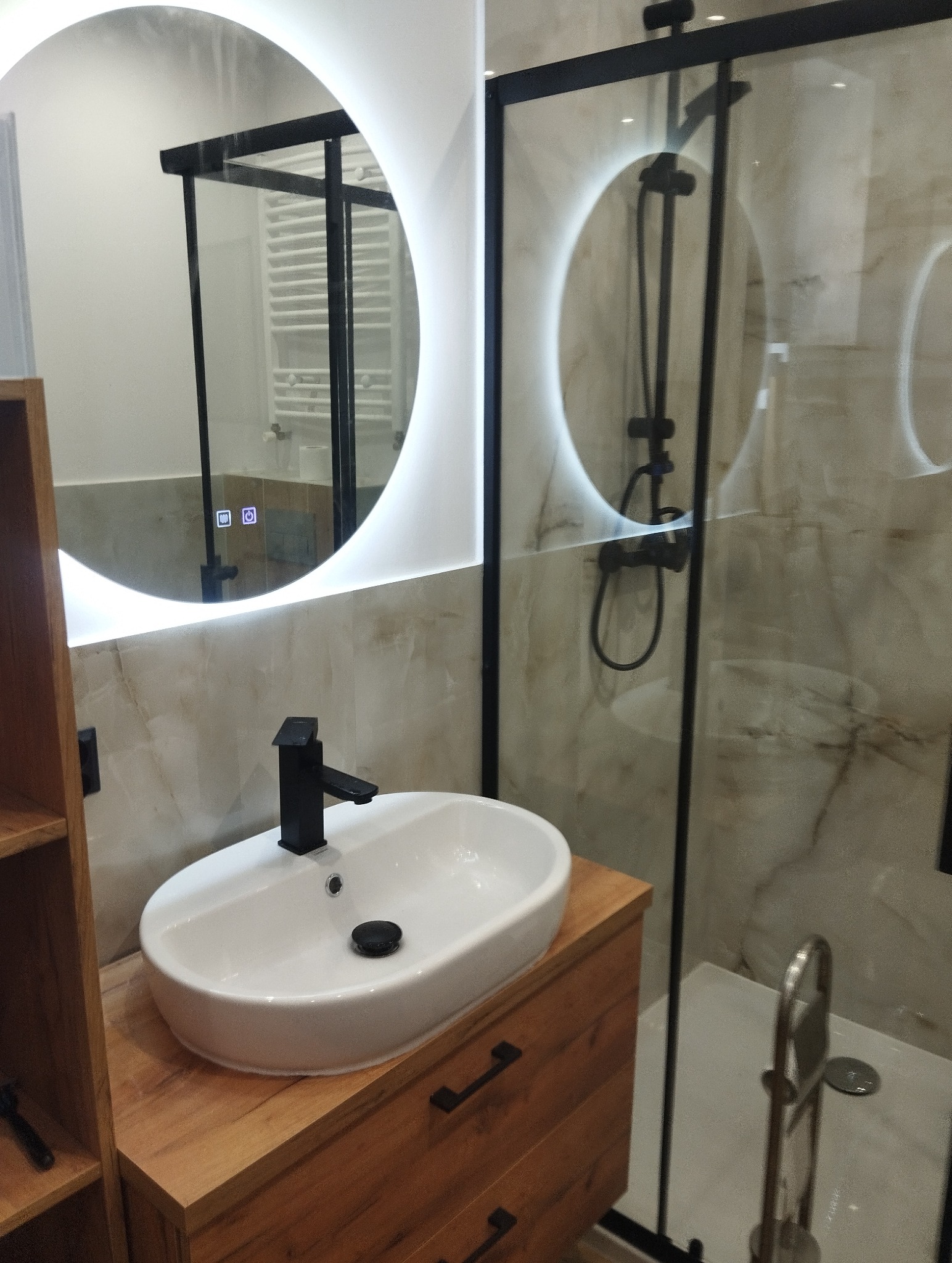 Nowoczesna łazienka z owalną umywalką na drewnianej szafce, czarną armaturą i podświetlanym lustrem. Kabina prysznicowa z czarnymi ramami i marmurowe płytki.