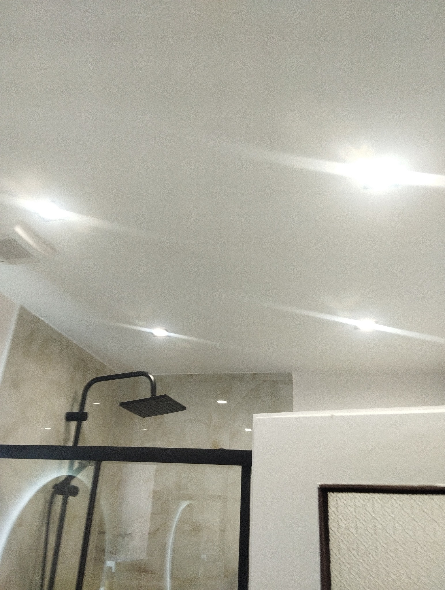 Nowoczesna kabina prysznicowa z czarną armaturą i szklaną ścianą, oświetlona punktowymi lampami sufitowymi. Minimalistyczny design łazienki.