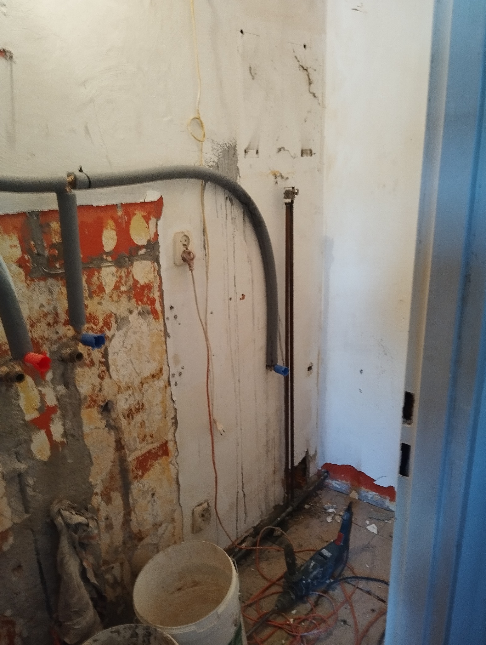 Wnętrze łazienki w trakcie remontu: odsłonięte rury, ściany z resztkami starej farby, wiadro i wiertarka na podłodze. Widoczne ślady po kuciu płytek.