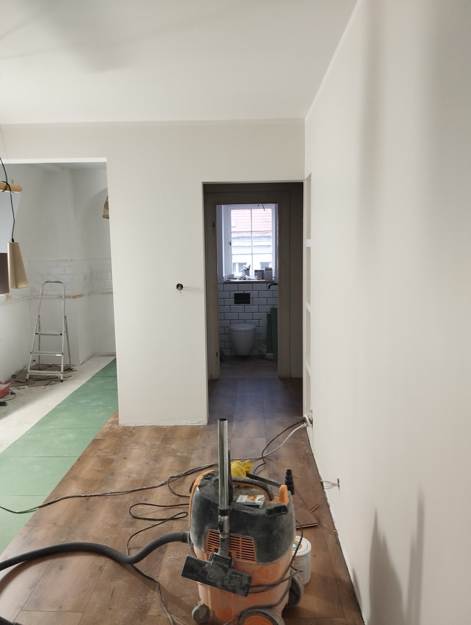 Wnętrze po malowaniu: białe ściany, podłoga z paneli, widok na wyremontowaną łazienkę z białymi płytkami. Na pierwszym planie odkurzacz przemysłowy.