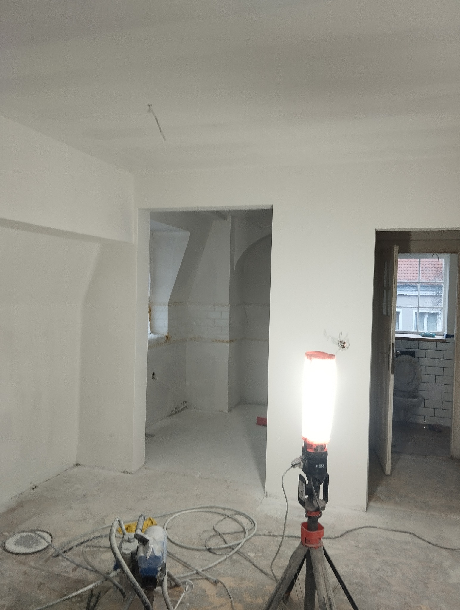 Wnętrze po szpachlowaniu i malowaniu na biało, widoczny agregat malarski i lampa budowlana. W tle wykończona łazienka z białymi płytkami i toaletą.