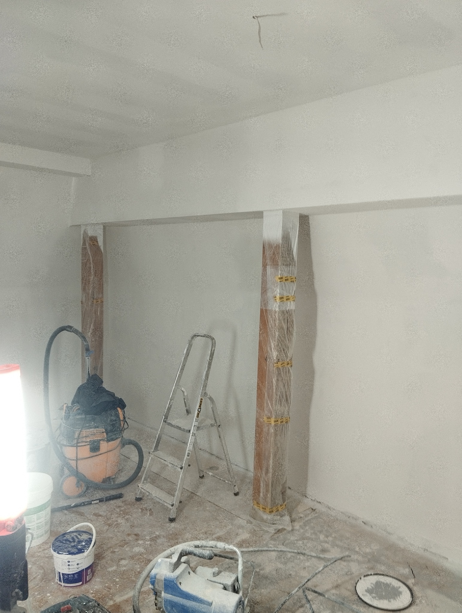 Przygotowanie do malowania: zabezpieczone folią kolumny, drabina, odkurzacz i narzędzia malarskie w pomieszczeniu z świeżo pomalowanymi ścianami na biało.